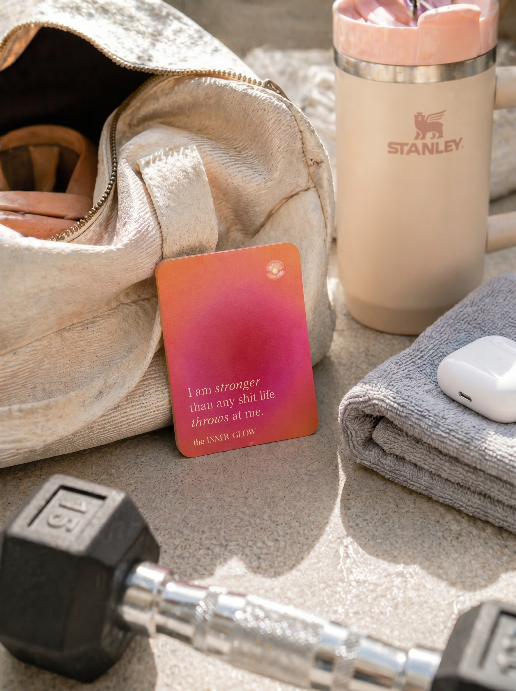 Stanley tumbler, dumbbell, towel, and motivational card on a sandy surface with affirmation card that says "I am stronger than any shit life throws at me"