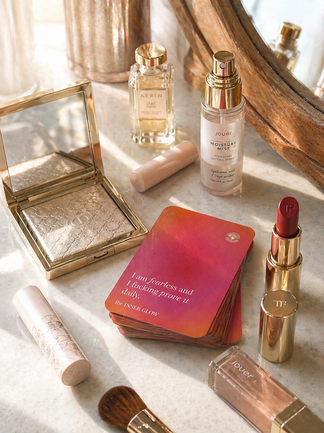 Collection of beauty products including a compact, lipstick, and perfume on a textured surface with deck of affirmation cards that says "I am fucking fearless and I fucking prove it daily"
