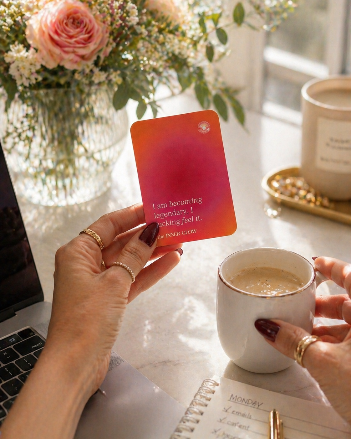 Person holding a card with text next to a cup of coffee on a desk with flowers and a laptop holding a bad ass affirmation card that says "I am becoming legendary and I can fucking feel it"